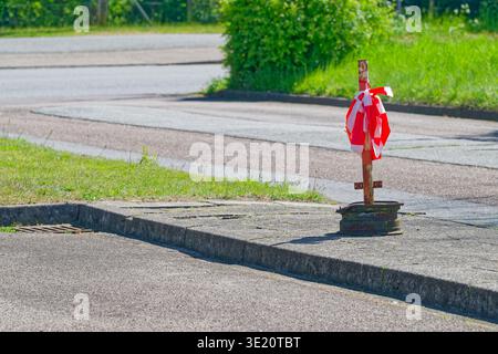 A rusty metal pole wrapped with red and white warning tape stands on a concrete sidewalk curb near a street with green grass and bushes in the backgro Stock Photo