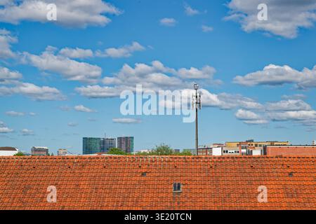 View of a city skyline featuring a red tiled roof in the foreground and a tall communication tower against a bright blue sky with scattered white clou Stock Photo
