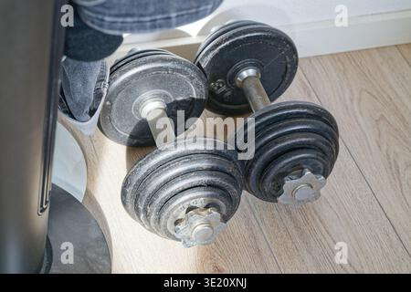 Two black adjustable dumbbells resting on a light wooden floor with natural light highlighting their textured metal weights and handles. Stock Photo