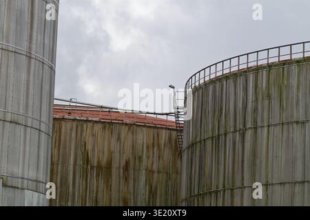 Three large cylindrical metal storage tanks with rust and moss stains, connected by a stairway and railing, set against a cloudy sky background. Stock Photo