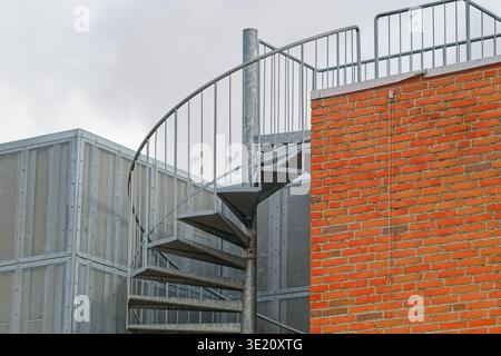 A metal spiral staircase with railings is attached to a red brick building with a perforated metal structure in the background under a cloudy sky. Stock Photo