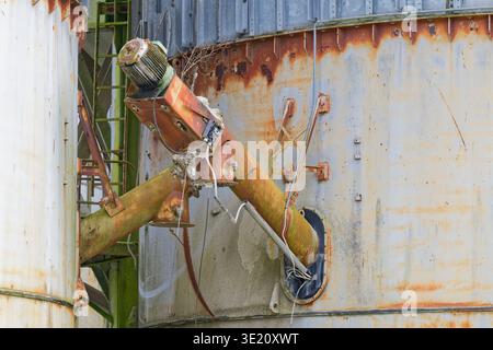 Old rusty industrial equipment with corroded metal pipes and electric motor attached to a weathered silo wall showing signs of decay and neglect. Stock Photo