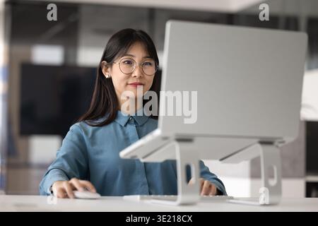 Focused Asian company worker woman in glasses working at laptop Stock Photo