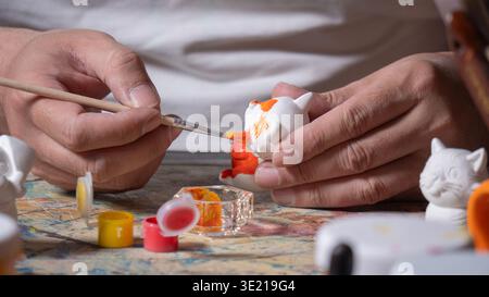 Close-up of a man's hands holding a fine brush with orange paint, painting a small white ceramic figure on a work table with paint jars. Stock Photo