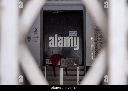 Broken glass door with padlock and red cloth. Metal gate frames the view. Stairs lead to the entrance. Signs and stickers adorn the wall. Dark, weathe Stock Photo
