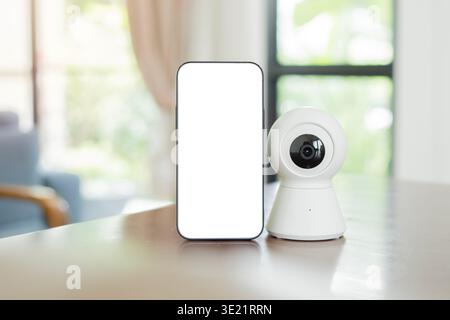A phone with blank screen setting next to a small surveillance camera on the table at home. Copy space, isolate screen. Stock Photo