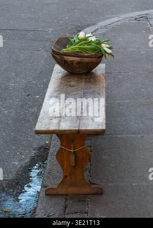 Rustic wooden bench on city sidewalk with a wooden bowl holding white tulips; quiet urban still life with copy space. Stock Photo