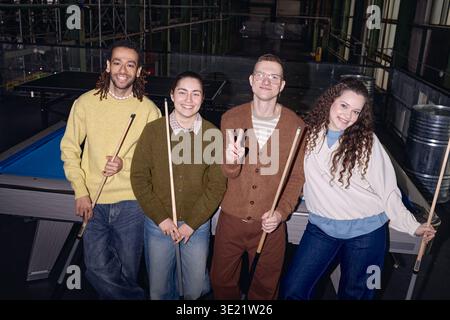 Diverse Young Adults Smiling and Posing with Pool Cues in Industrial Setting Stock Photo