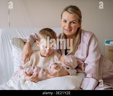 Mother holding newborn baby while sitting on a hospital bed with her older son beside her in a hospital room after childbirth. Tender family moment Stock Photo