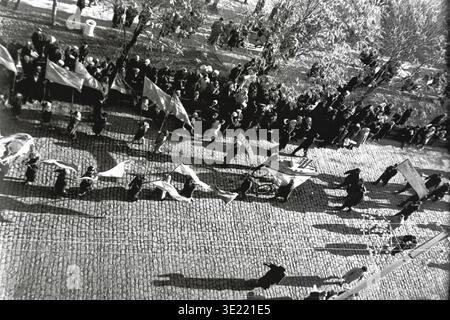 Fascinating black and white archival photograph from 1967 showing a top-down view of the November 7 demonstration moving along a textured cobblestone street in the city center of Slavyansk. The marching column casts long, dramatic shadows in the bright sunlight, with participants carrying waving flags and a creative decorative float on wheels. This dynamic street photography preserves the memory of creative civic participation, historical city infrastructure, and the nostalgic atmosphere of a peaceful Donbas before the war. Stock Photo