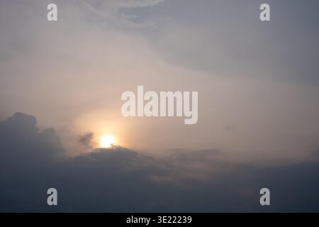 Sun peeks through clouds at dusk. Soft light glows behind dark, swirling clouds. Sky blends gray to warm hues. Natural, moody atmosphere captured. Clo Stock Photo