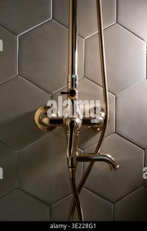 Shower fixture mounted on hexagonal tiled wall. Gold-toned spout and hose connected to wall mount. Modern bathroom design with soft ambient lighting Stock Photo