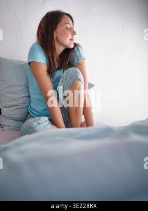 Woman in blue pajamas sitting on bed against a white wall, looking relaxed and thoughtful. Germany Stock Photo