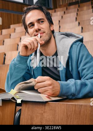 Young man in hoodie sitting in lecture hall with a pen and open book, looking thoughtful. Germany Stock Photo