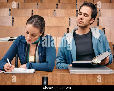 Two young adults in a lecture hall, one taking notes and the other reading a book.   Germany Stock Photo