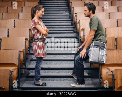 Two young adults conversing on empty lecture hall steps with wooden seats. Germany Stock Photo