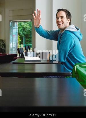 A smiling man in a blue hoodie sits at a table waving in a bright, modern cafÈ. Germany Stock Photo