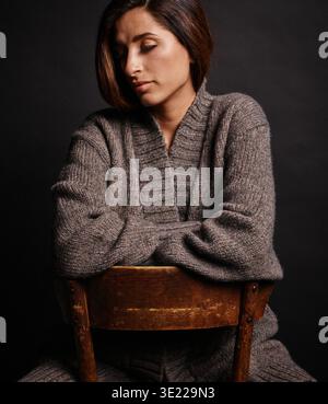 Woman in a cozy knitted sweater sitting on a wooden chair with a dark background Germany Stock Photo