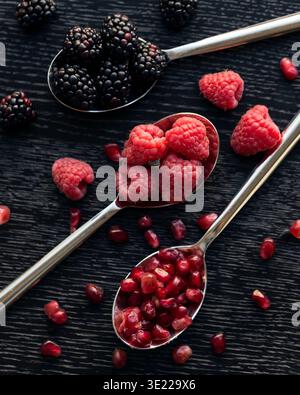 Three spoons filled with blackberries, raspberries, and pomegranate seeds on a dark wooden surface. Stock Photo