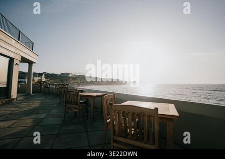 Outdoor cafe seating overlooks calm ocean. Wooden tables and chairs sit empty, bathed in soft morning light. Coastal cliffs rise behind tranquil water Stock Photo