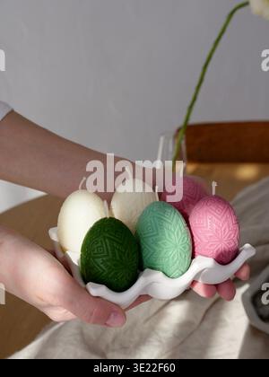 Female hands holding a ceramic tray with colorful beeswax candles in the shape of decorated Easter eggs. Spring holiday home decor. Close-up. Stock Photo