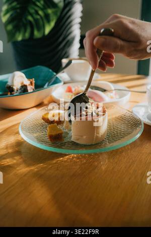 Hand dips spoon into creamy dessert. Golden light spills across wooden table. Spoon lifts a bite, sugared and rich. Dessert sits on textured glass pla Stock Photo