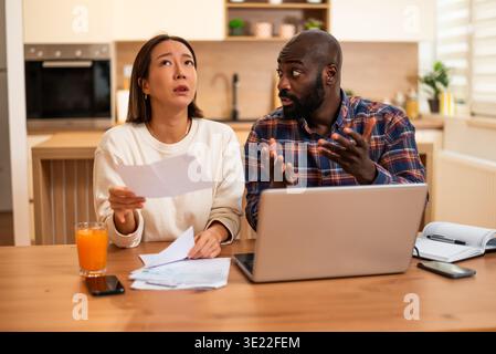 A couple sits at a table reviewing bills and documents, looking concerned as they compare paperwork with information on a laptop. Stock Photo