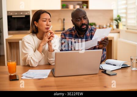 A couple sits at a table reviewing bills and documents, looking concerned as they compare paperwork with information on a laptop. Stock Photo
