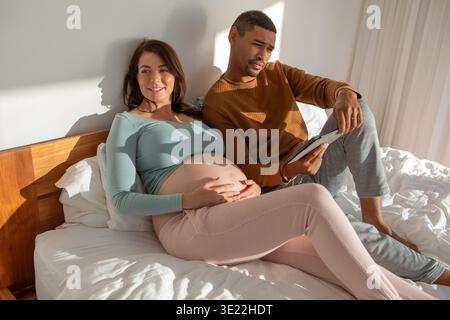 Diverse female partners sitting on home bed wooden headboard pillows cradling belly holding tablet. Pregnancy, intimacy, relaxation, home, wellness, nurturing, connection || Model released Stock Photo