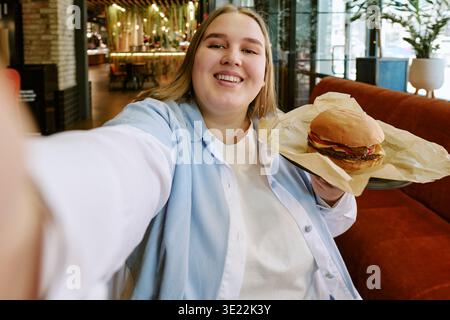 Portrait of Young Adult Woman Smiling While Holding Hamburger Indoors Stock Photo