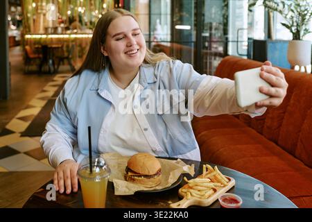 Young Woman Taking Selfie While Eating Burger and French Fries Indoors Stock Photo