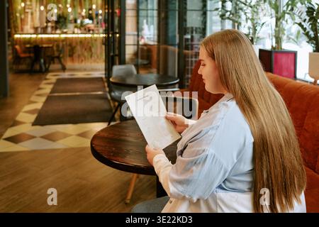 Young Woman Sitting Indoors Reading Menu at Restaurant Table Stock Photo