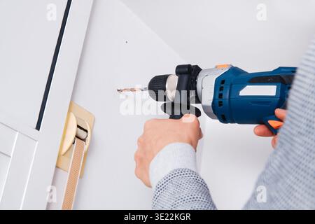 a man drills a hole in a white wall with a blue electric drill, preparing the surface for installing a metal bracket that will support a curtain rod Stock Photo