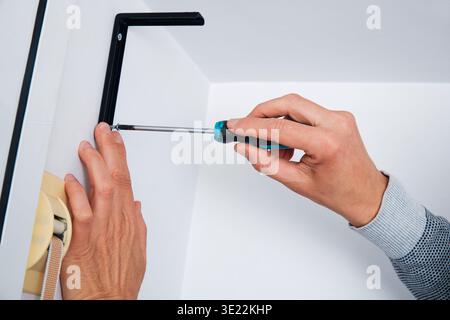 a man tightens a screw with a screwdriver to secure a black metal bracket on a white wall, holding the bracket steady with his other hand Stock Photo