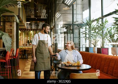 Young Woman Paying Waiter with Smartphone in Modern Restaurant Stock Photo
