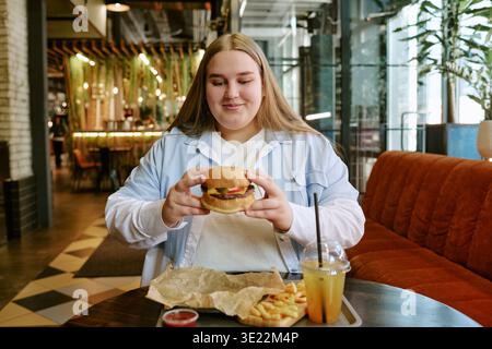 Young Woman Smiling While Holding Burger and Sitting in Cafe Stock Photo