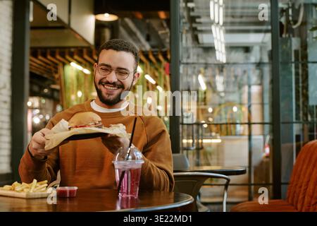 Young Adult Man Smiling While Eating Burger and French Fries Indoors Stock Photo