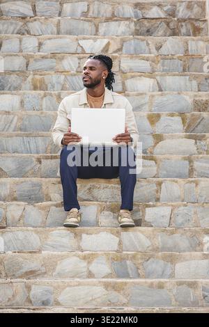 A man sits nonchalantly on some stone steps, holding a white computer. He seems pensive, enjoying a quiet moment outdoors in the daylight. The setting Stock Photo