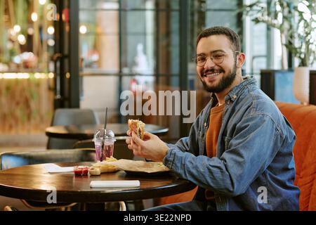 Portrait of Young Adult Man Smiling While Eating Burger Indoors Stock Photo