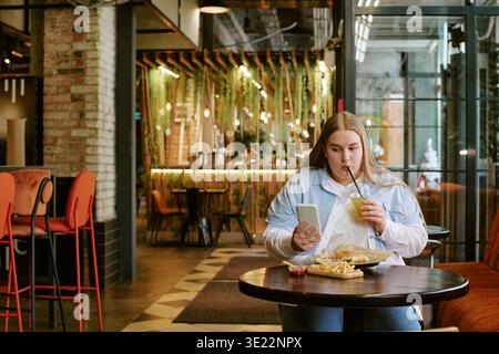 Young Adult Woman Sitting in Cafe Using Smartphone While Eating Lunch Stock Photo