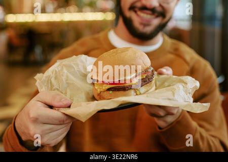 Young Adult Man Smiling While Holding Cheeseburger Indoors Stock Photo
