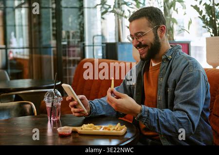 Young Adult Black Man Smiling While Using Smartphone and Eating French Fries Stock Photo