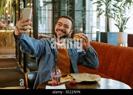 Young Adult Man Taking Selfie While Eating Burger in Restaurant Stock Photo