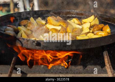 Potatoes cooking over open flame outdoors. Close-up of sliced potatoes frying in a large pan over an open fire, with steam, flames, and rustic outdoor Stock Photo