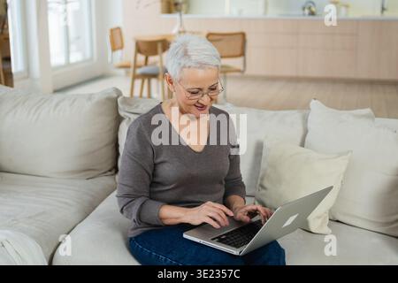 Confident stylish happy middle aged senior woman using laptop at home. Stylish older mature 60s grandmother sitting at couch looking at computer screen typing chatting reading writing email Stock Photo