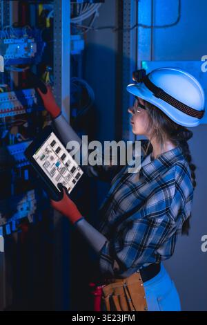 Female electrician inspecting electrical panel with tablet in server room. Power engineer. Stock Photo