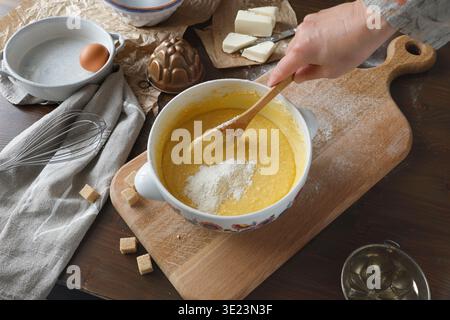 Mixing flour into cake batter in ceramic bowl on wooden board. Close-up of hand with wooden spoon preparing dough with ingredients like eggs and Stock Photo