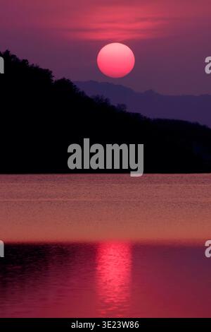 A dramatic crimson sunset over a calm lake with a glowing red sun reflecting on the water, framed by dark mountain silhouettes. The scene creates a po Stock Photo