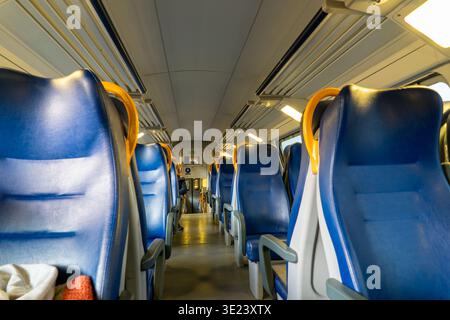 The perspective shot shows rows of empty blue passenger seats with yellow grab handles in a clean train compartment, illuminated by overhead lights Stock Photo