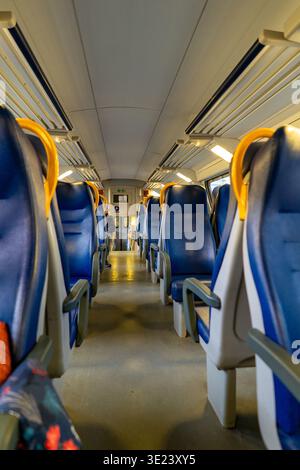 The perspective shot shows rows of empty blue seats with yellow grab handles inside a passenger train carriage, illuminated by overhead lights Stock Photo
